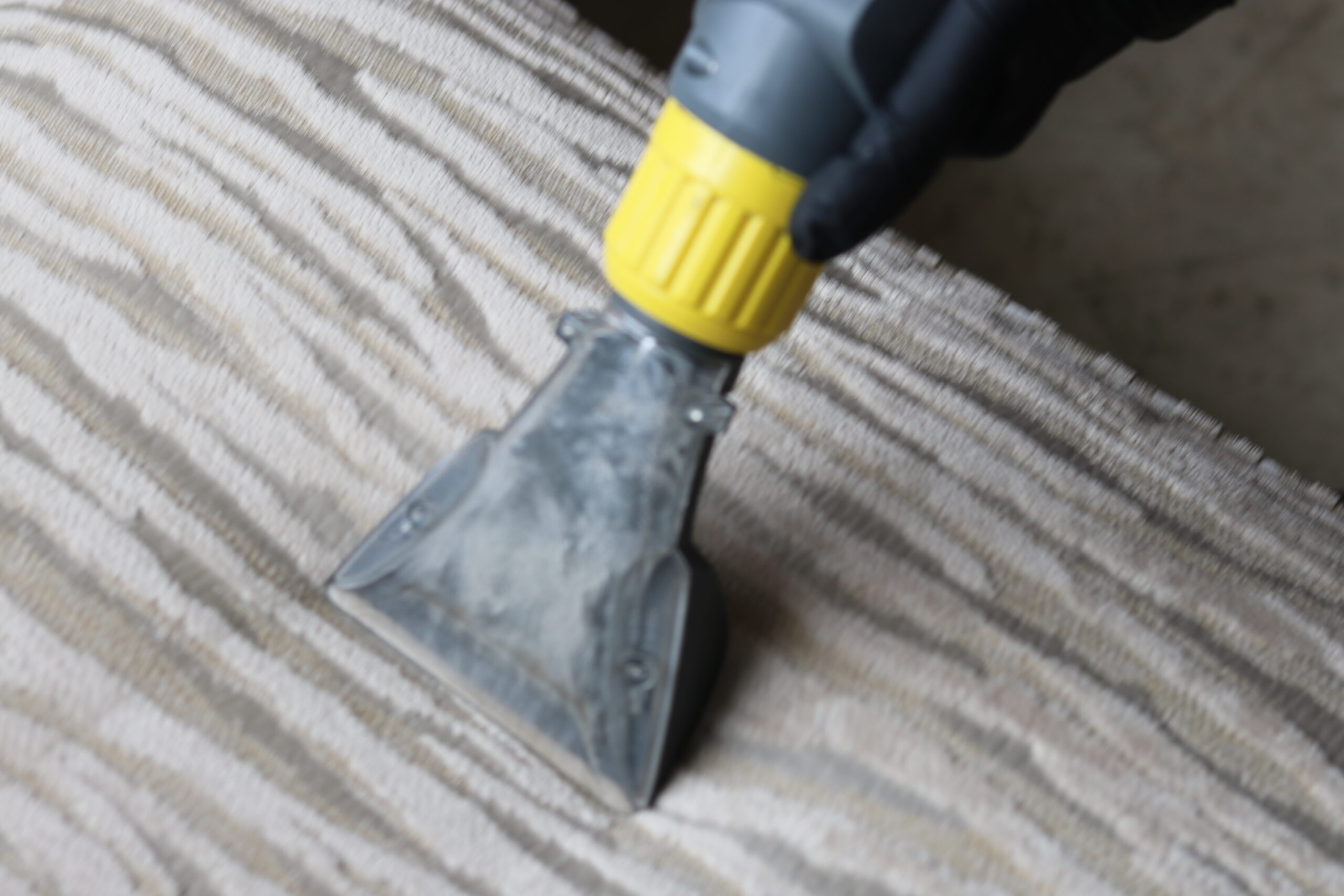 A professional cleaner in uniform deep cleaning a beige fabric sofa using a vacuum and shampooing tool.
